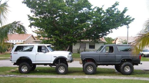 1995 Ford Bronco - Two lifted Ford Broncos are parked on a street in front of houses and trees.