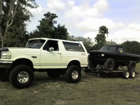 1995 Ford Bronco - A white SUV is parked next to a black pickup truck that is loaded onto a trailer.