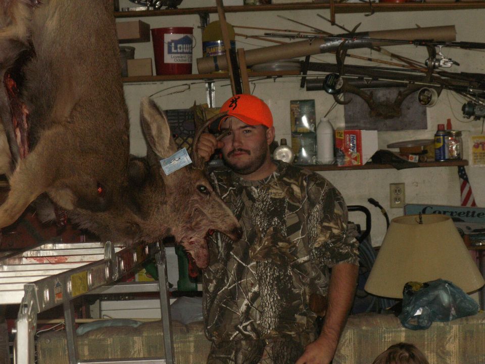 Man in camouflage holding a harvested deer, with shelves of hunting gear and supplies in the background