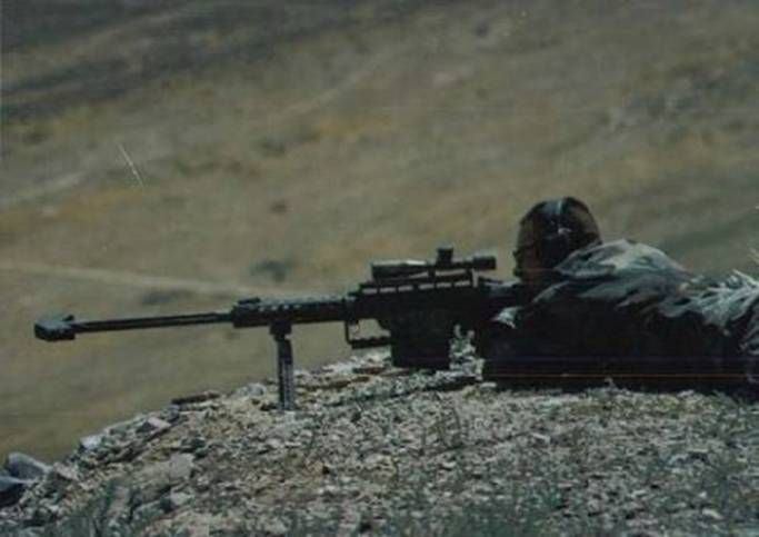 Person in camouflage prone behind a large scoped rifle on a rocky hillside