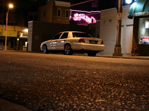 2003 Ford Crown Victoria - A white 2003 Ford Crown Victoria is parked on a city street at night, illuminated by streetlights and neon signs.