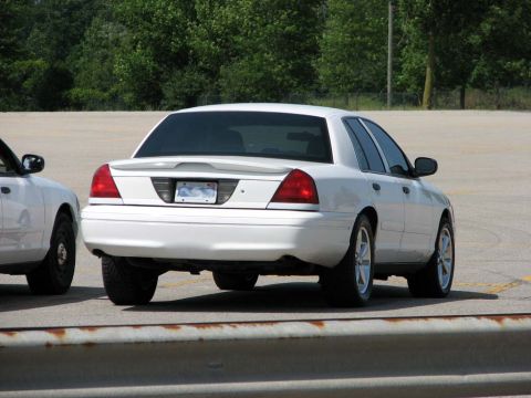 2003 Ford Crown Victoria - A white 2003 Ford Crown Victoria is parked next to another vehicle, showing its rear end and tinted windows.
