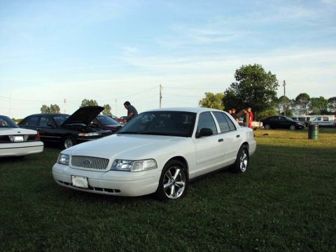 2003 Ford Crown Victoria - A white 2003 Ford Crown Victoria is parked on a grassy field with other vehicles in the background.