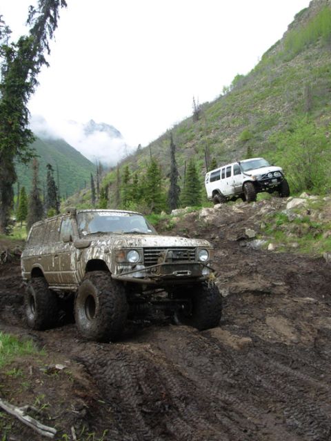 2000 Ford E350 - A mud-caked tan Toyota Land Cruiser wagon on huge tractor-lug tires sits in deep rutted mud on a mountain trail, wearing a front bumper wit…