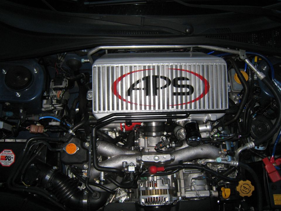 Engine bay with a large silver intercooler labeled "APS" in red oval. Blue hoses and black wiring are visible