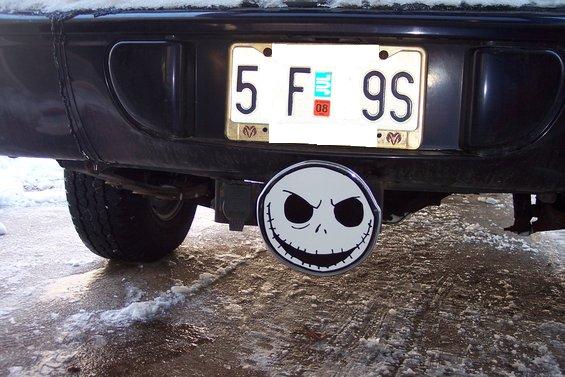 Rear of a dark-colored vehicle with a license plate and a Jack Skellington hitch cover. Snow is on the ground