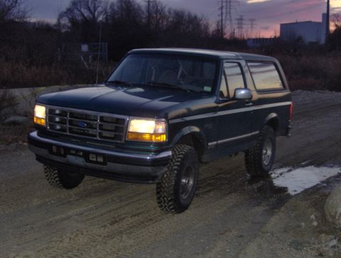 1996 Ford Bronco - A dark green Ford Bronco with lifted suspension is seen driving on a dirt road at dusk.