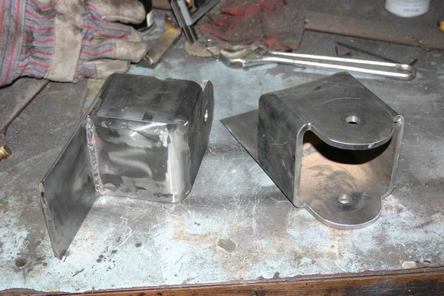 Two metal brackets, one cube-shaped with a side plate, the other U-shaped with mounting holes, sit on a workbench