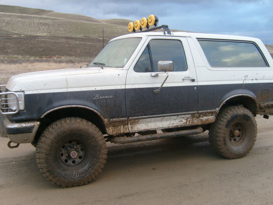 White and blue lifted Ford Bronco, side view, muddy off-road tires and roof light bar