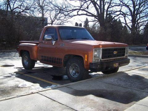 1979 Chevrolet C/K Pickup - A classic orange pickup truck is parked in a lot with bare trees in the background.