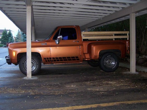 Orange pickup truck parked under a carport, side view. It has a wooden bed insert and black checkered decals on the door
