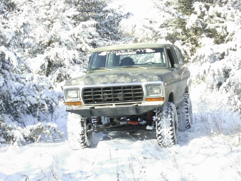 1978 Ford Bronco with lifted suspension and off-road tires driving through a snowy wooded area