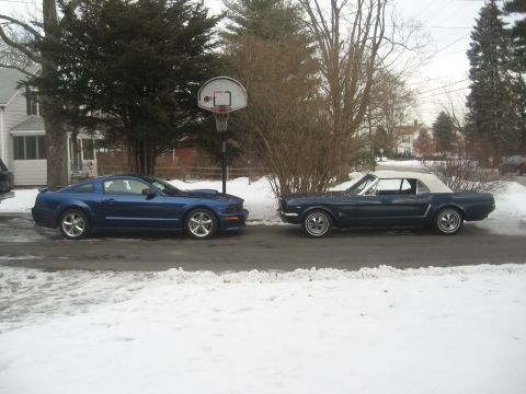 2007 Ford Mustang - Two Ford Mustangs, a modern coupe and a classic convertible, are parked side-by-side on a street with snow on the ground.