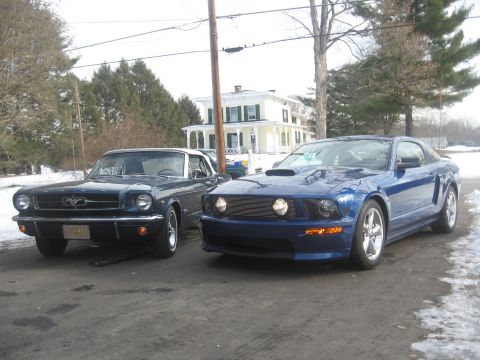2007 Ford Mustang - Two Ford Mustangs, a classic convertible and a modern coupe, are parked side-by-side on a street with snow on the ground.