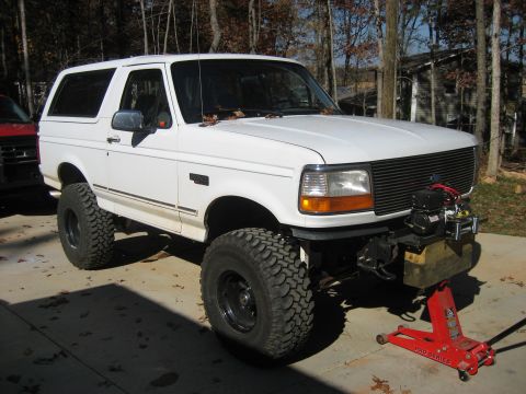 1996 Ford Bronco - Lifted white two-door Ford Bronco shot from the front three-quarter angle on a concrete driveway in a wooded yard, wearing large mud-terrai…