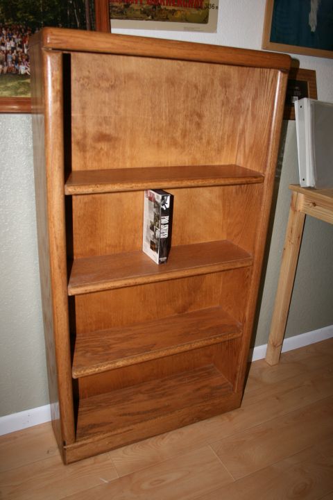 2003 Jeep TJ - A wooden bookshelf with three shelves stands against a wall, holding a single book on its middle shelf.