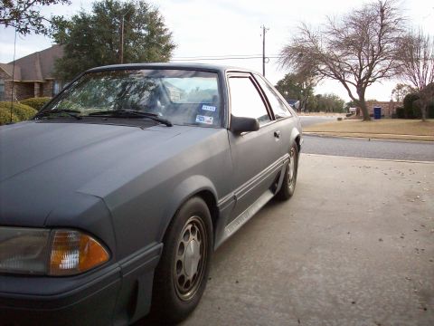 1989 Ford Mustang - A gray Ford Mustang coupe is seen from a three-quarter front angle, parked in a driveway with a house and trees in the background.