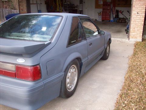 1989 Ford Mustang - The rear quarter panel and taillight of a gray Ford Mustang are visible, parked in a garage driveway.
