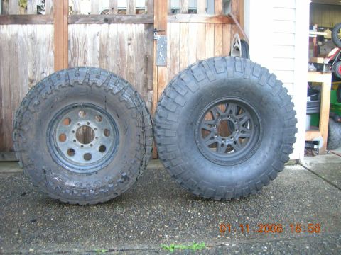 1996 Ford Bronco - A pair of aggressive off-road tires and wheels are displayed side-by-side in front of a wooden gate.