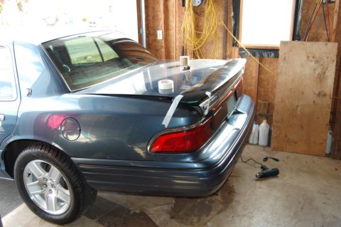 1985 Mercury Grand Marquis - A spoiler is being attached to the trunk of a car in a garage with tape.