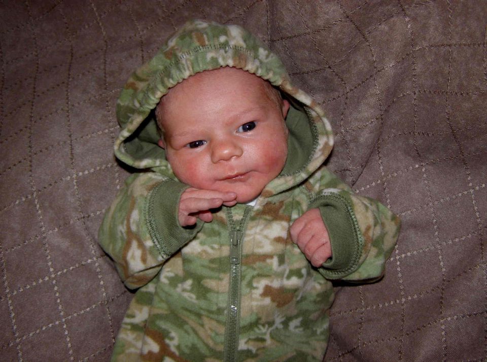 Newborn baby in a camouflage hooded outfit on a quilt