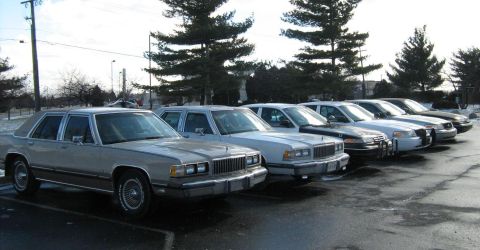 1988 Mercury Grand Marquis - A line of tan and white sedans, including a 1988 Mercury Grand Marquis, parked in a lot on a cloudy day.