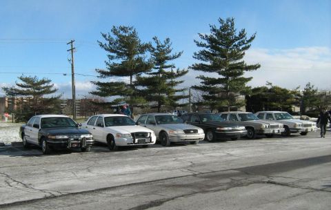 1988 Mercury Grand Marquis - A line of sedans, likely police or security vehicles, are parked in a lot under a bright sky.