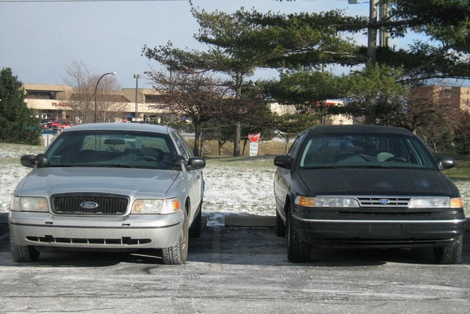 Silver sedan and black sedan parked front-end out, snow on ground, trees and buildings in background