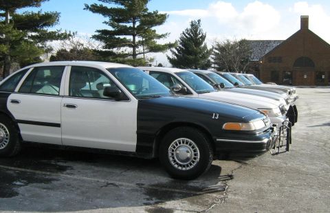 1988 Mercury Grand Marquis - Line of black-and-white Ford Crown Victoria Police Interceptor sedans parked in a lot, lead car marked number 13, brick building behind.