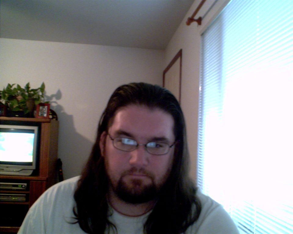 Man with long dark hair and glasses looking to the side, with a television and plant in the background