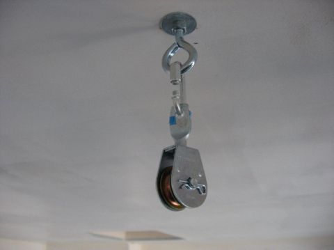 1978 Ford Bronco - A small zinc-plated rope pulley with a bronze-colored sheave hangs from a white ceiling, suspended by a snap hook clipped to an eye bolt an…