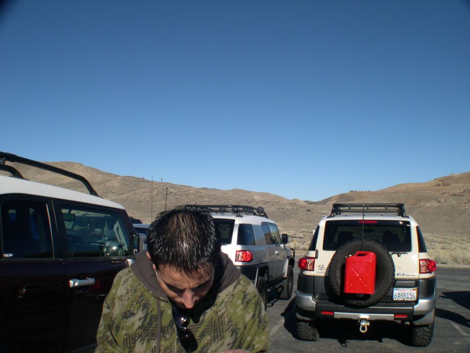 Two Toyota FJ Cruisers parked, one with a red gas can on the spare tire. Man in camo jacket in front