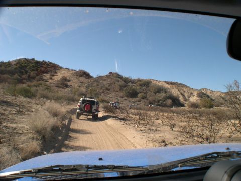 2007 Toyota FJ Cruiser - Multiple Toyota FJ Cruisers driving on a dirt road through a dry, hilly terrain under a clear blue sky.