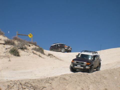2007 Toyota FJ Cruiser - Two Toyota FJ Cruisers navigate a sandy incline under a clear blue sky, with a directional sign visible on the left.