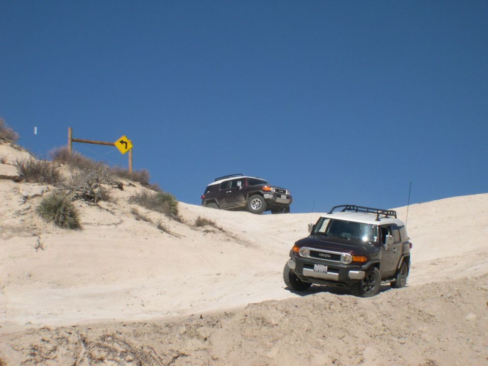 Two dark-colored 2007 Toyota FJ Cruisers driving on a sandy hill with a blue sky