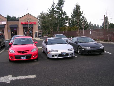2007 Mazda 3 - A red 2007 Mazda 3 is parked in a lot alongside two Mitsubishi Eclipses, with a "Bad Ass Coffee" store in the background.