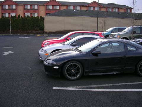 2007 Mazda 3 - Three cars are parked in a lot outside a hotel. The closest car is black, followed by a silver car and a red car.