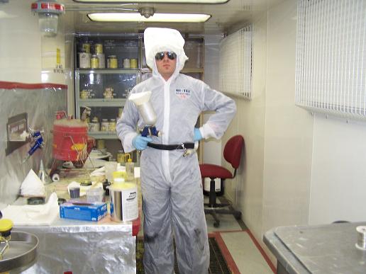 Person in a white protective suit and hood, wearing sunglasses, holding a spray gun. They are standing in a room with shelves of supplies and a red drum