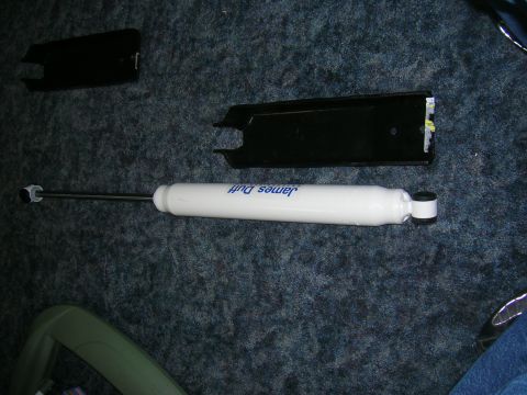 1979 Ford Bronco - white James Duff branded shock absorber lies on dark carpet next to two black metal mounting bracket pieces.