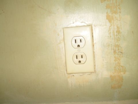1993 Ford Bronco - A cream-colored duplex electrical outlet with a matching wall plate mounted on a rough, pale green wall showing patched and scraped areas.