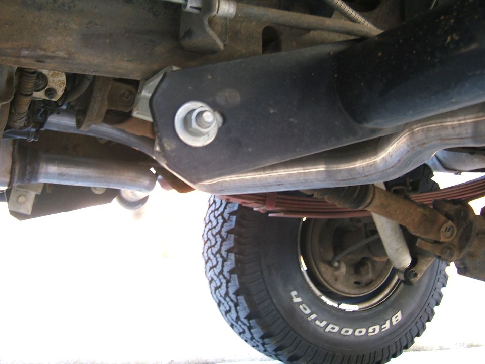 Underside view of a vehicle's rear axle, leaf springs, and exhaust pipe. Tire visible