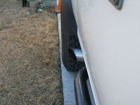 1991 Ford Bronco - A close-up side view shows the exhaust pipe and tire of a white vehicle parked on a concrete surface next to dry grass.