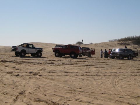 1996 Ford Bronco - Several off-road vehicles are gathered on sandy terrain under a clear blue sky.