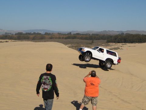 1996 Ford Bronco - A white SUV is captured mid-air as it jumps over a sand dune, with two onlookers observing the stunt.