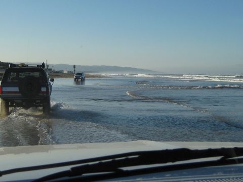 1996 Ford Bronco - Two SUVs drive along the wet sand near the ocean waves under a clear blue sky.