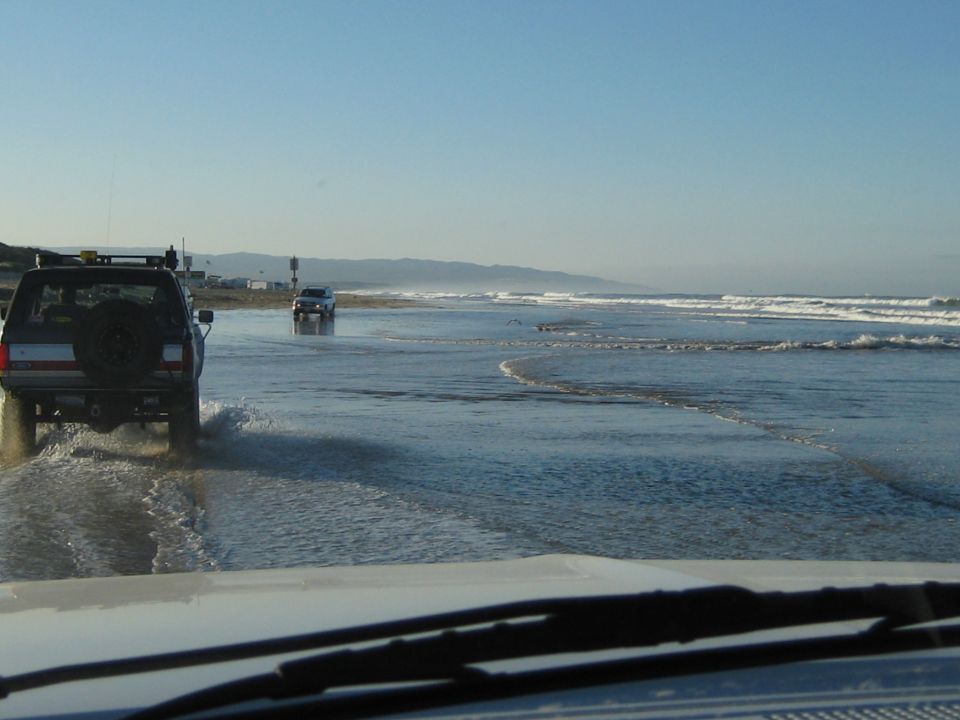 Rear view of a 1996 Ford Bronco driving on a wet beach with another SUV in the distance