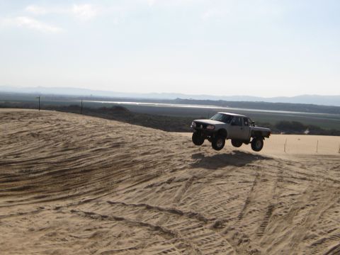 1996 Ford Bronco - A lifted pickup truck catches air while cresting a sand dune on a sunny day.