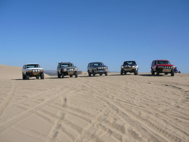 Five off-road SUVs parked on sand dunes under a clear blue sky