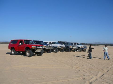 1988 Ford Bronco - A group of Ford Broncos and other SUVs are lined up on a sandy landscape under a clear blue sky.