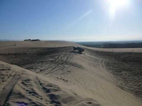 1988 Ford Bronco - A dark SUV navigates a sandy incline under a bright, sunny sky, leaving tire tracks in its wake.